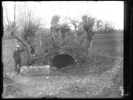 Un homme en costume posant à côté d'un [sarcophage ?], devant l'entrée d'un souterrain, dans un champ.